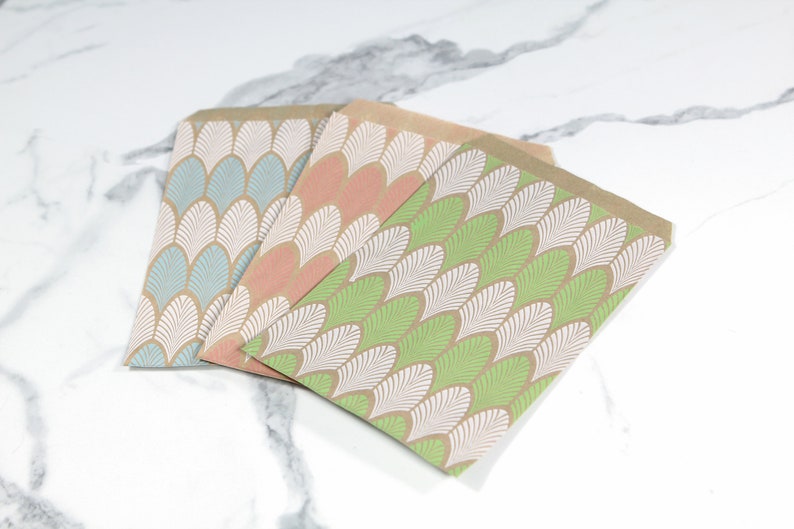 K&ouml;nnte beinhalten: Drei Papier-Geschenkbeutel mit braunem Hintergrund und einem wei&szlig;en und gr&uuml;nen geometrischen Muster. Die Beutel sind &uuml;bereinander gestapelt, wobei der gr&uuml;ne Beutel oben, der rosa Beutel in der Mitte und der blaue Beutel unten liegt.