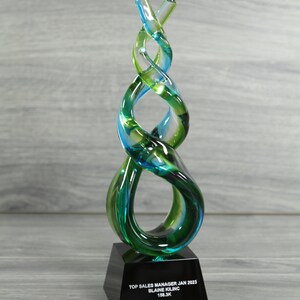May include: A glass sculpture award with a blue and green swirl design on a black base. The base has the text "TOP SALES MANAGER JAN 2023 BLAINE KILINC 158.3K"