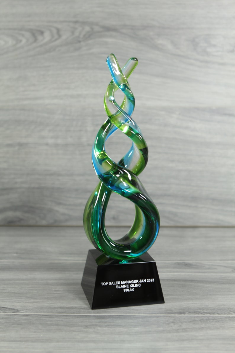May include: A glass sculpture award with a blue and green swirl design on a black base. The base has the text "TOP SALES MANAGER JAN 2023 BLAINE KILINC 158.3K"