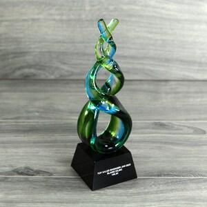 May include: A green and blue glass award with a black base. The base has the text "Top Sales Manager Jan 2023 Blake Sterling Team".