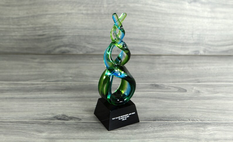May include: A green and blue glass award with a black base. The base has the text "Top Sales Manager Jan 2023 Blake Sterling Team".