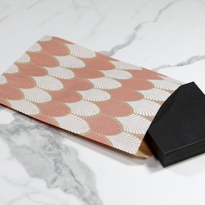 K&ouml;nnte beinhalten: Eine schwarze Geschenkbox mit einem rosa-wei&szlig;en, gemusterten Papierumschlag. Der Umschlag hat ein geometrisches Muster aus &uuml;berlappenden Schuppen.