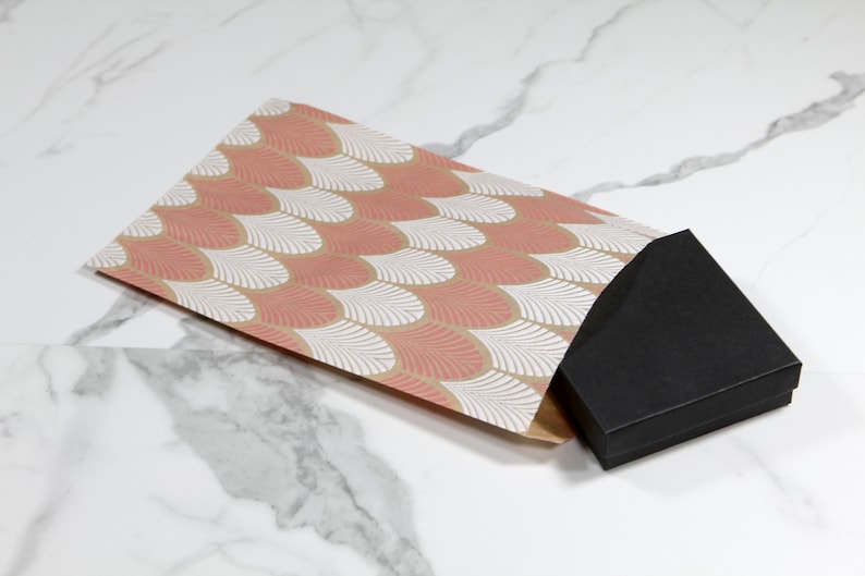 K&ouml;nnte beinhalten: Eine schwarze Geschenkbox mit einem rosa-wei&szlig;en, gemusterten Papierumschlag. Der Umschlag hat ein geometrisches Muster aus &uuml;berlappenden Schuppen.