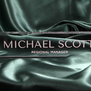 May include: A clear acrylic desk nameplate with the text "MICHAEL SCOTT" and "REGIONAL MANAGER" in white lettering.