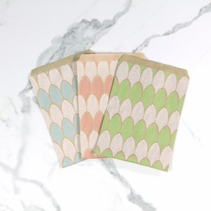 K&ouml;nnte beinhalten: Drei Papier-Geschenkbeutel mit einem wei&szlig;en und gr&uuml;nen geometrischen Muster. Die Beutel sind gefaltet und &uuml;bereinander gestapelt. Der obere Beutel ist gr&uuml;n, der mittlere Beutel ist rosa und der untere Beutel ist blau.