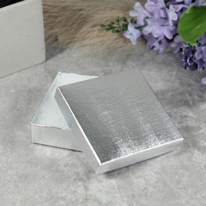 May include: A silver gift box with a textured lid is open, revealing a white interior. The box is sitting on a gray surface.