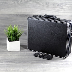 May include: A black leather briefcase with a white stitched border and a black strap. The briefcase is closed and sitting on a wood surface.