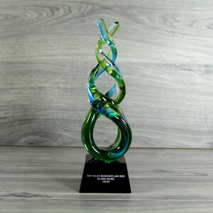May include: A green and blue glass award with a black base. The base has the text "Top Sales Manager Jan 2023, Blaine Kline, 118.3%".