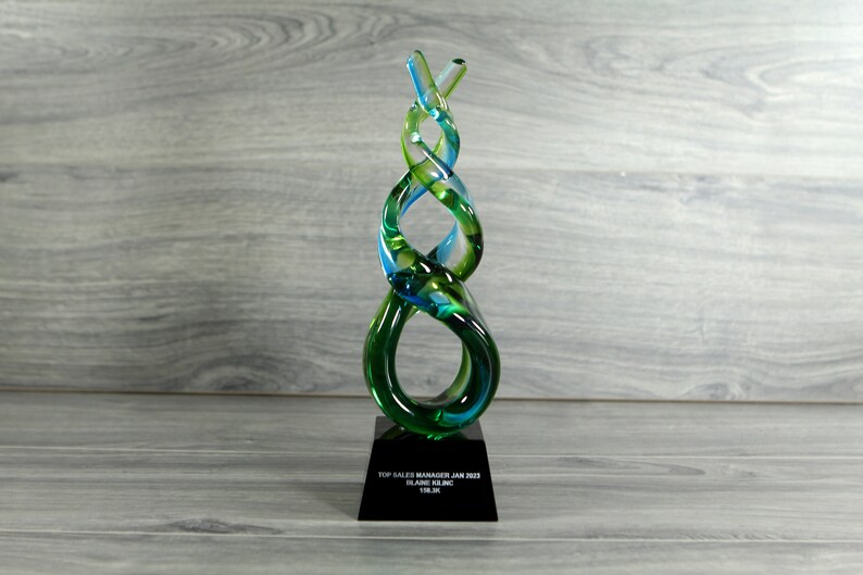 May include: A green and blue glass award with a black base. The base has the text "Top Sales Manager Jan 2023, Blaine Kline, 118.3%".