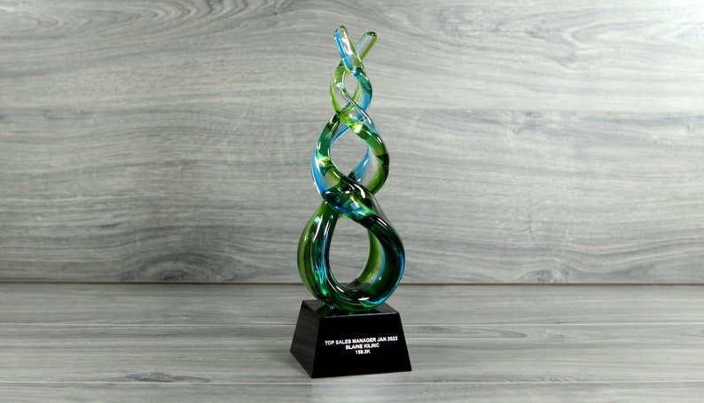 May include: A blue and green glass award with a black base. The base has the text "TOP SALES MANAGER JAN 2023 BLAINE SLINC 18.3K"