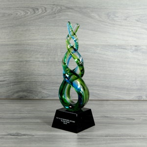 May include: A glass award trophy with a swirling, abstract design in shades of green and blue. The base is black and features the text "Top Sales Manager June 2022, Blaine Island, Team".