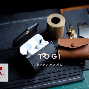 Peut inclure: Un étui en cuir noir avec un bouton doré et le texte "TOGI handmade" pour AirPods. L'étui est ouvert et montre des AirPods blancs à l'intérieur. L'étui est sur une surface noire avec un étui en cuir marron et d'autres outils de maroquinerie.