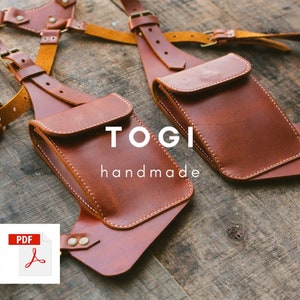 May include: Two brown leather camera straps with a pocket on each strap. The straps are connected by a harness. The text "TOGI handmade" is printed on the straps.