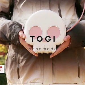 Pode incluir: Uma bolsa branca em forma de caveira com cavidades oculares rosa e uma alça rosa. O texto "TOGI handmade" está impresso na bolsa. A bolsa é segurada por uma pessoa que usa um casaco.