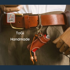 Op de afbeelding: Een bruine leren sleutelhanger met een rode aansteker erin. De sleutelhanger is bevestigd aan een bruine leren riem met een messing gesp. De tekst "ToGi Handmade" is zichtbaar op de afbeelding.