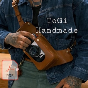 May include: A brown leather camera strap with a brown leather camera case attached. The strap is worn over the shoulder of a person wearing a blue denim jacket. The text "ToGi Handmade" is visible on the jacket.
