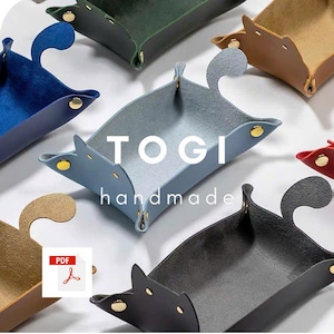 Könnte beinhalten: Eine Auswahl handgefertigter Ledertabletts in verschiedenen Farben, darunter Blau, Grün und Grau. Jedes Tablett hat ein einzigartiges Katzenohrendesign und ist mit goldfarbenen Nieten befestigt. Die Wörter "TOGI" und "handmade" sind aufgedruckt.