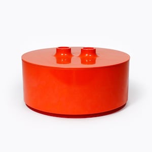 May include: A bright orange, cylindrical container with a flat, circular lid. Two small, cylindrical features protrude from the lid's surface. The container has a smooth, glossy finish and is set against a white backdrop. The design is modern and minimalist.