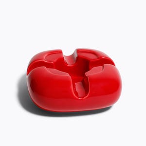 Peut inclure: Un cendrier rouge vif de forme carrée arrondie. Il comporte quatre encoches incurvées pour poser les cigarettes et un puits central. Le design est minimaliste et contemporain, avec une surface lisse et réfléchissante.