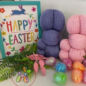Puede incluir: Tres decoraciones de conejito de Pascua de punto en morado, rosa y turquesa. Los conejitos están hechos de hilo y tienen una textura esponjosa. Los conejitos están sentados en una mesa con un cartel que dice "Feliz Pascua" en letras de colores.