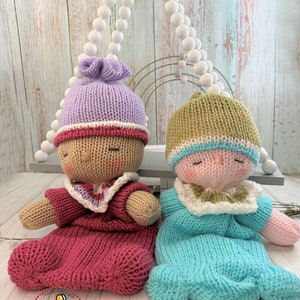 May include: Two knitted dolls wearing sleep sacks. The doll on the left is wearing a purple hat and a red sleep sack. The doll on the right is wearing a green hat and a blue sleep sack.