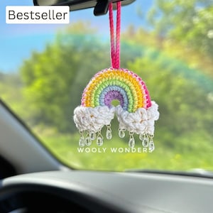 May include: A handmade crochet rainbow car charm with a colorful arched design, white cloud accents, and dangling clear beads. The charm is suspended from a pink cord and features the text "WOOLY WONDERS". The word "Bestseller" is also visible.