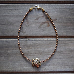May include: A black and gold beaded anklet with a gold turtle charm. The anklet is on a wooden surface.