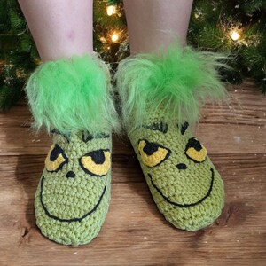 May include: A pair of green crocheted slippers with a Grinch face design. The slippers have yellow eyes, a black smile, and green fluffy trim at the ankles. The slippers are on a wooden surface.