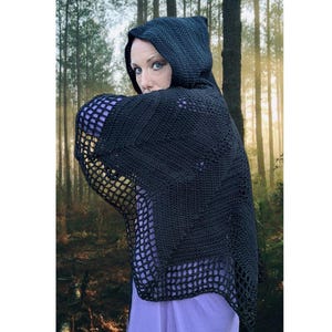Puede incluir: Un chal con capucha negro de ganchillo con un diseño geométrico. El chal tiene una capucha y un patrón de red en los bordes inferiores. El fondo es un bosque con la luz del sol filtrándose entre los árboles.
