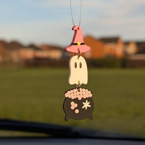 May include: A pink witch hat, a white ghost, and a black cauldron with pink and white stars hang from a rearview mirror.
