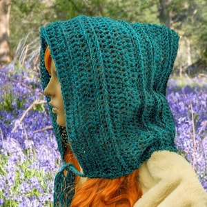 May include: A teal crocheted hooded scarf with a fringe. The scarf is worn by a person with long red hair. The person is standing in a field of bluebells.