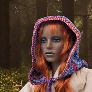May include: A woman with long, bright orange hair wears a crocheted hooded scarf with a colorful pattern of blue, pink, and purple. The scarf is tied at the neck and the hood is pulled up over her head. She is wearing a white fur coat.