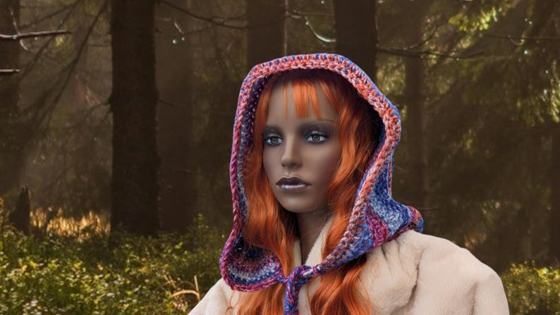 May include: A woman with long, bright orange hair wears a crocheted hooded scarf with a colorful pattern of blue, pink, and purple. The scarf is tied at the neck and the hood is pulled up over her head. She is wearing a white fur coat.