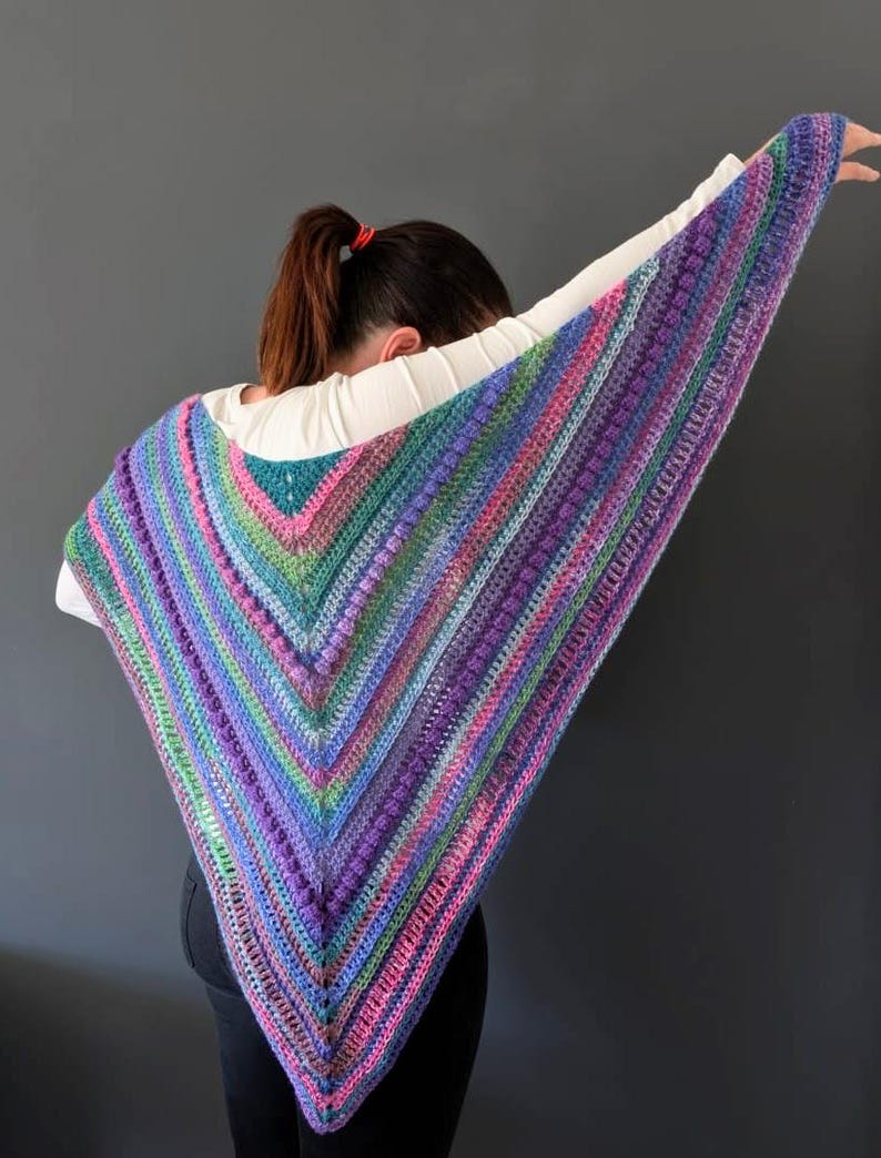 Pu&ograve; includere: Uno scialle all'uncinetto fatto a mano, di forma triangolare e multicolore. Lo scialle presenta una vivace miscela di colori, tra cui viola, blu, verde e rosa, disposti a righe. La trama sembra avere un punto in rilievo, tipo a pallini.