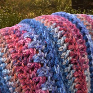 May include: A crocheted blanket with stripes of pink, blue, and white yarn. The blanket is folded in half and has a soft, textured look.