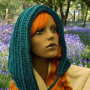 May include: A teal blue crocheted hooded scarf with a long, flowing design. The scarf is worn by a person with long, bright orange hair. The person is standing in a field of blue flowers.
