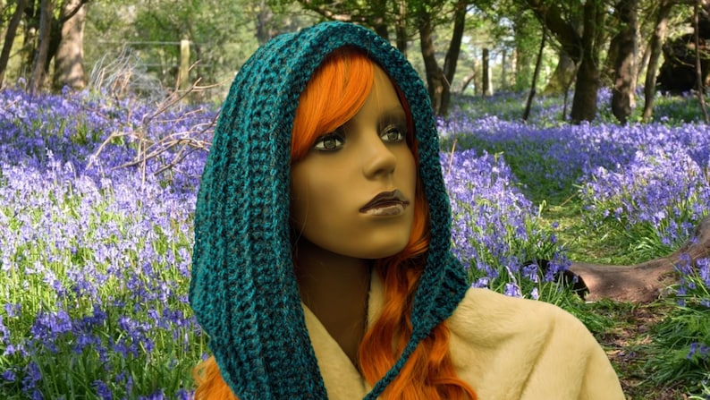 May include: A teal blue crocheted hooded scarf with a long, flowing design. The scarf is worn by a person with long, bright orange hair. The person is standing in a field of blue flowers.
