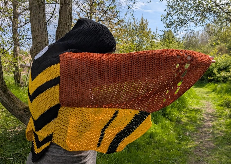 May include: A handmade crochet bee-themed hooded shawl. The shawl features a black hood, yellow and black striped body, and brown wings with cut-out details. The garment is displayed outdoors with a natural background.