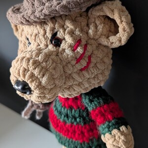 May include: Crocheted plush figure of a bear-like character with a brown hat and a striped red and green sweater. The bear has red stitched scars on its face and a black nose. The plush is made of a soft, textured yarn.
