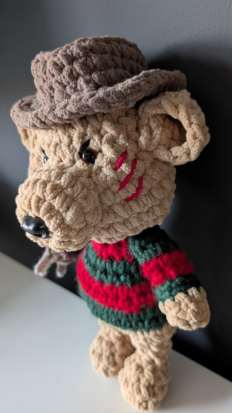 May include: Crocheted plush figure of a bear-like character with a brown hat and a striped red and green sweater. The bear has red stitched scars on its face and a black nose. The plush is made of a soft, textured yarn.