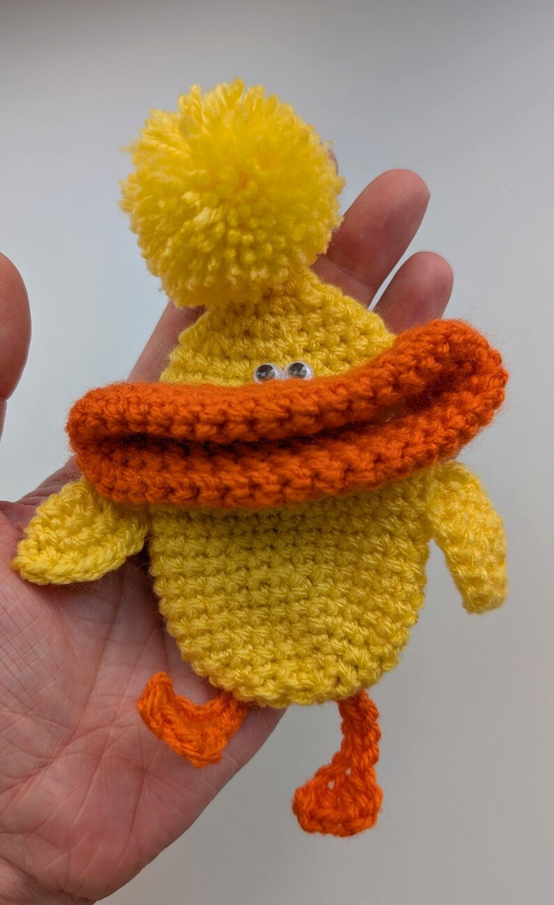 May include: A handmade yellow crochet duck with an orange beak and feet. The duck has a yellow pom-pom on its head and googly eyes. This whimsical item is a charming addition to any collection.