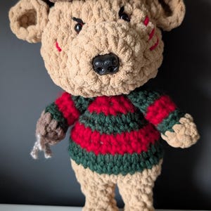May include: A handmade crochet bear doll wearing a brown hat and a striped red and green sweater. The bear has a black nose, angry eyes, and red slashes on its face. The bear's claws are silver.