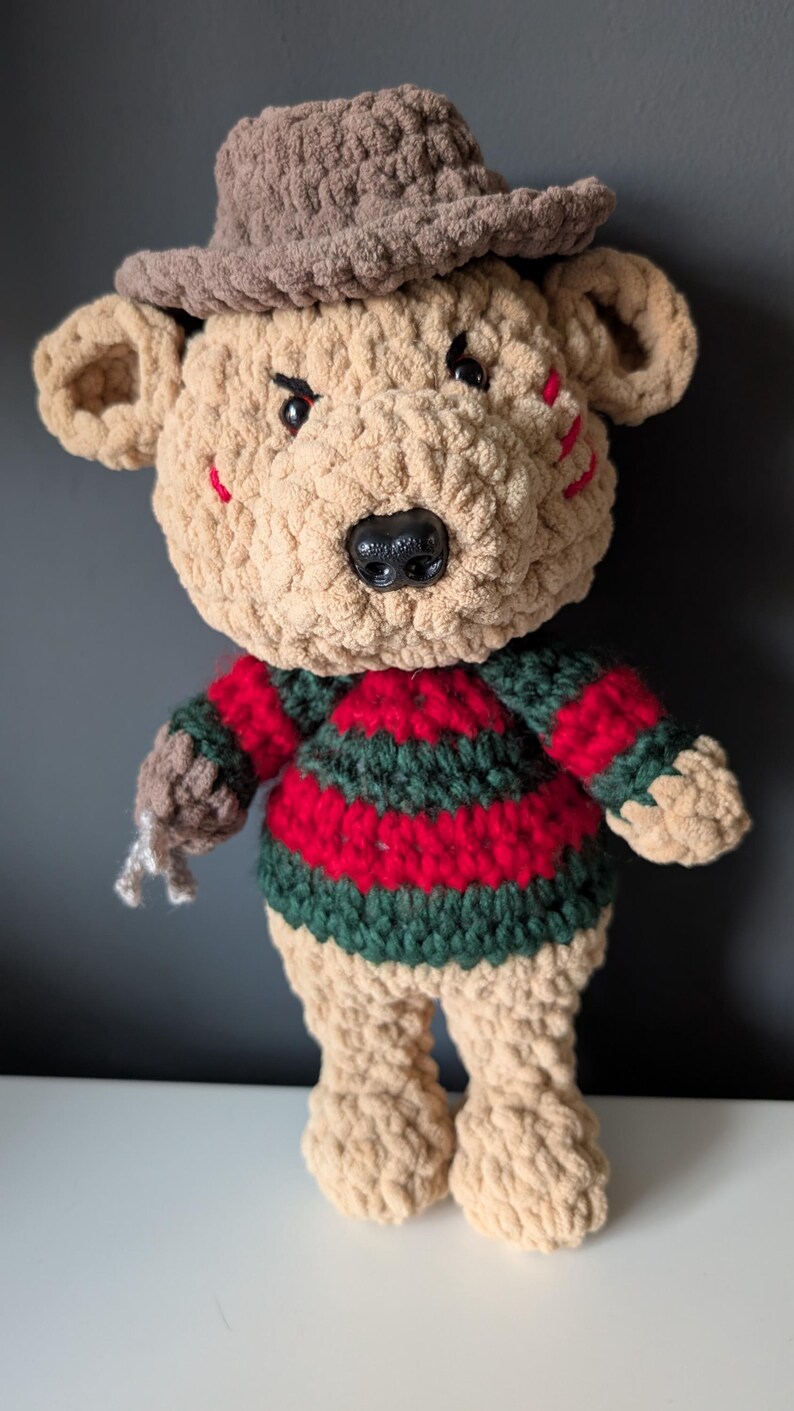 May include: A handmade crochet bear doll wearing a brown hat and a striped red and green sweater. The bear has a black nose, angry eyes, and red slashes on its face. The bear's claws are silver.