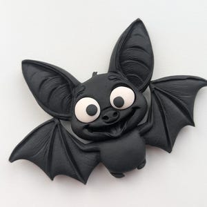 May include: A black cartoon bat with large, expressive eyes and a friendly smile. The bat has large ears and detailed wings, with a light-colored area around the eyes. The bat is a Halloween or gothic-themed item.