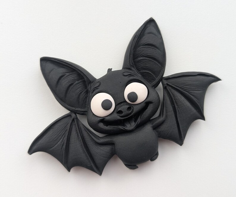 May include: A black cartoon bat with large, expressive eyes and a friendly smile. The bat has large ears and detailed wings, with a light-colored area around the eyes. The bat is a Halloween or gothic-themed item.