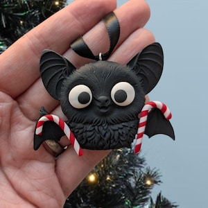 Puede incluir: Adorno navide&ntilde;o de murci&eacute;lago negro con grandes ojos blancos y una sonrisa. El murci&eacute;lago sostiene dos bastones de caramelo a rayas rojas y blancas. Se adjunta una cinta negra para colgarlo. Decoraci&oacute;n festiva.