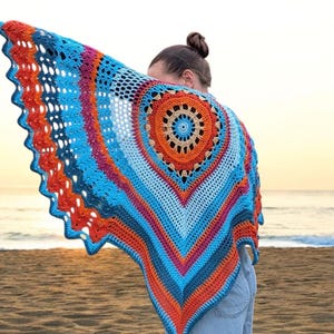 Op de afbeelding: Een kleurrijke, gehaakte sjaal met een cirkelvormig ontwerp in blauw, oranje en roze tinten. De sjaal wordt gedragen, met een strand en de zee op de achtergrond.