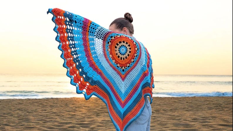 Op de afbeelding: Een kleurrijke, gehaakte sjaal met een cirkelvormig ontwerp in blauw, oranje en roze tinten. De sjaal wordt gedragen, met een strand en de zee op de achtergrond.