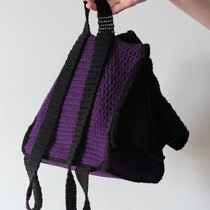 May include: A handmade crochet backpack in a striking color combination. The bag features a deep purple body with black straps and trim. The straps are adjustable and the bag is held by a hand against a white background.