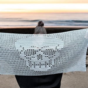 May include: A light blue, open-weave crochet shawl with a skull design. The shawl is held up against a beach backdrop, with the ocean and sky visible in the background. The crochet pattern is visible.